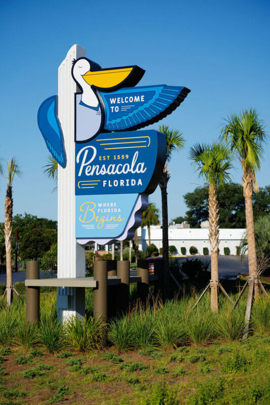 A daytime shot of the "Welcome to Pensacola, Florida" sign with a pelican, set against a clear blue sky.