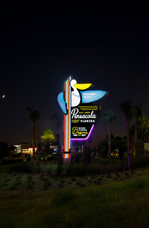 A vertical shot of the "Welcome to Pensacola, Florida" sign at night, illuminated by neon lights, with a crescent moon in the dark sky.