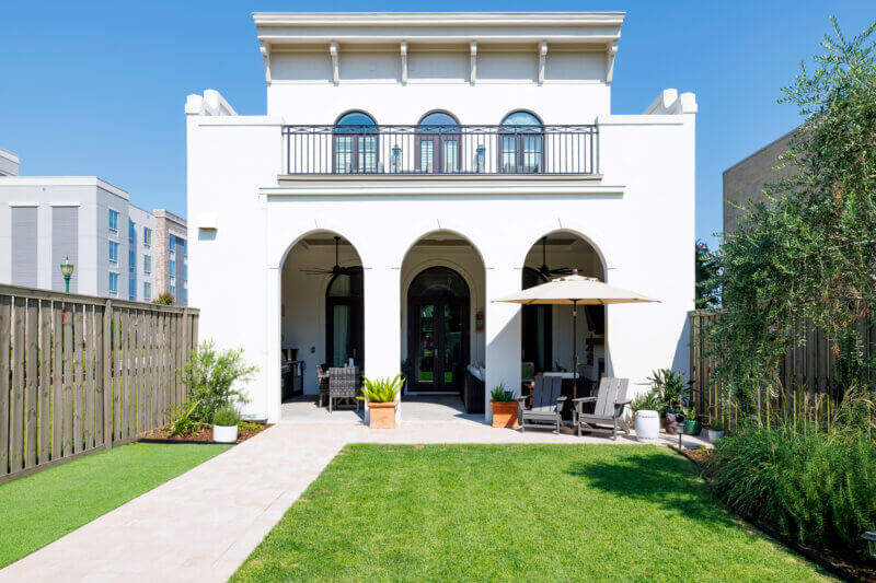 The back exterior of a two-story white stucco home with three arched doorways on the ground floor and a balcony on the second floor, looking out over a green backyard with a stone path.