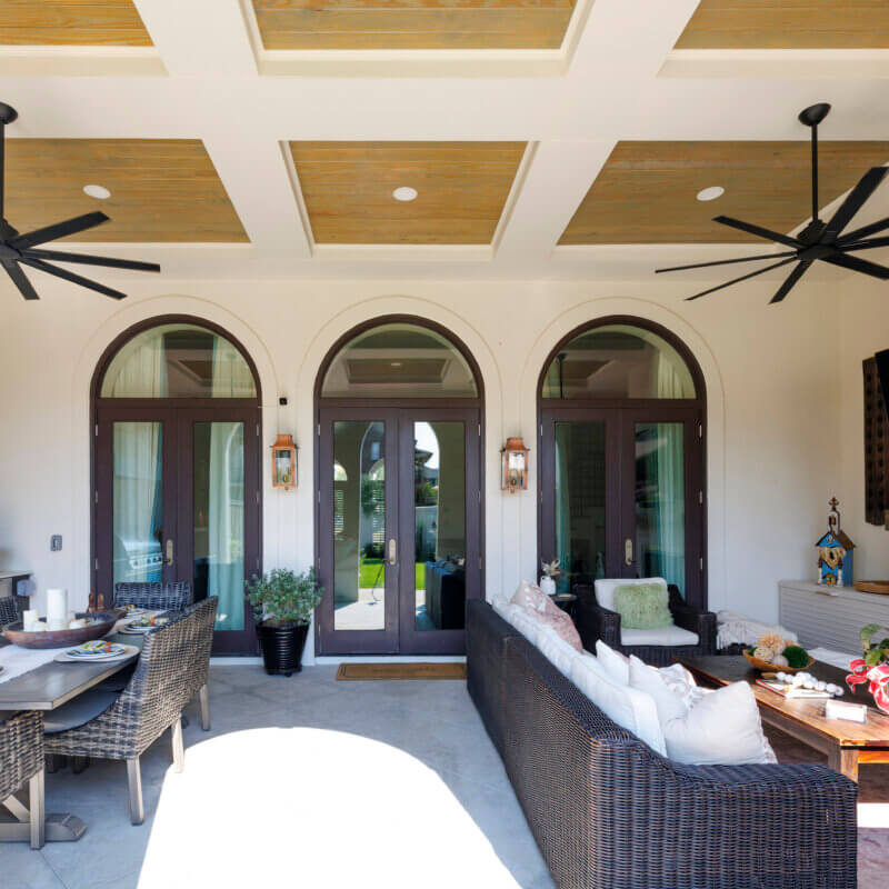 A covered outdoor patio with a dining table and a long sofa, featuring two large arched entryways and a detailed wood-paneled ceiling.