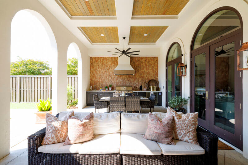 An outdoor living and dining area with a long wicker sofa, a dining table, and a full outdoor kitchen against a unique patterned backsplash wall.