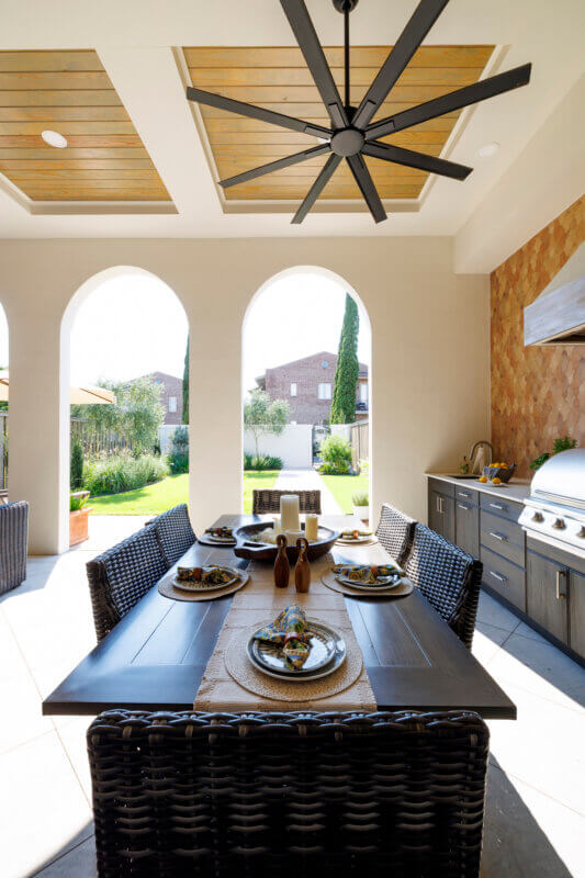 An outdoor dining table with place settings and a large ceiling fan, situated on a covered patio with three arched openings to the backyard.