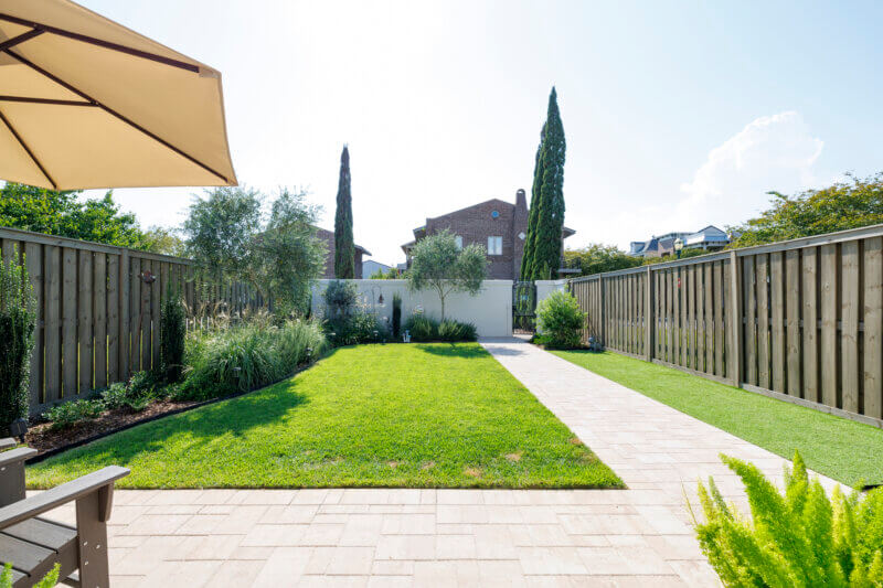 A shot of a backyard with a neatly manicured lawn, a stone paver pathway, and various plants and trees.