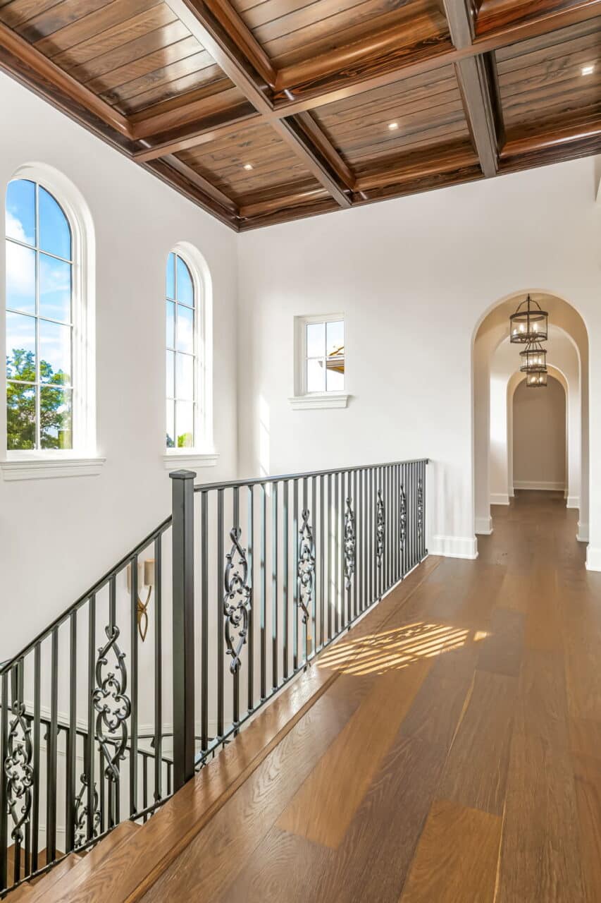 Upper-level hallway featuring dark wood floors, an ornate iron stair railing, large arched windows, and a dark wood coffered ceiling.
