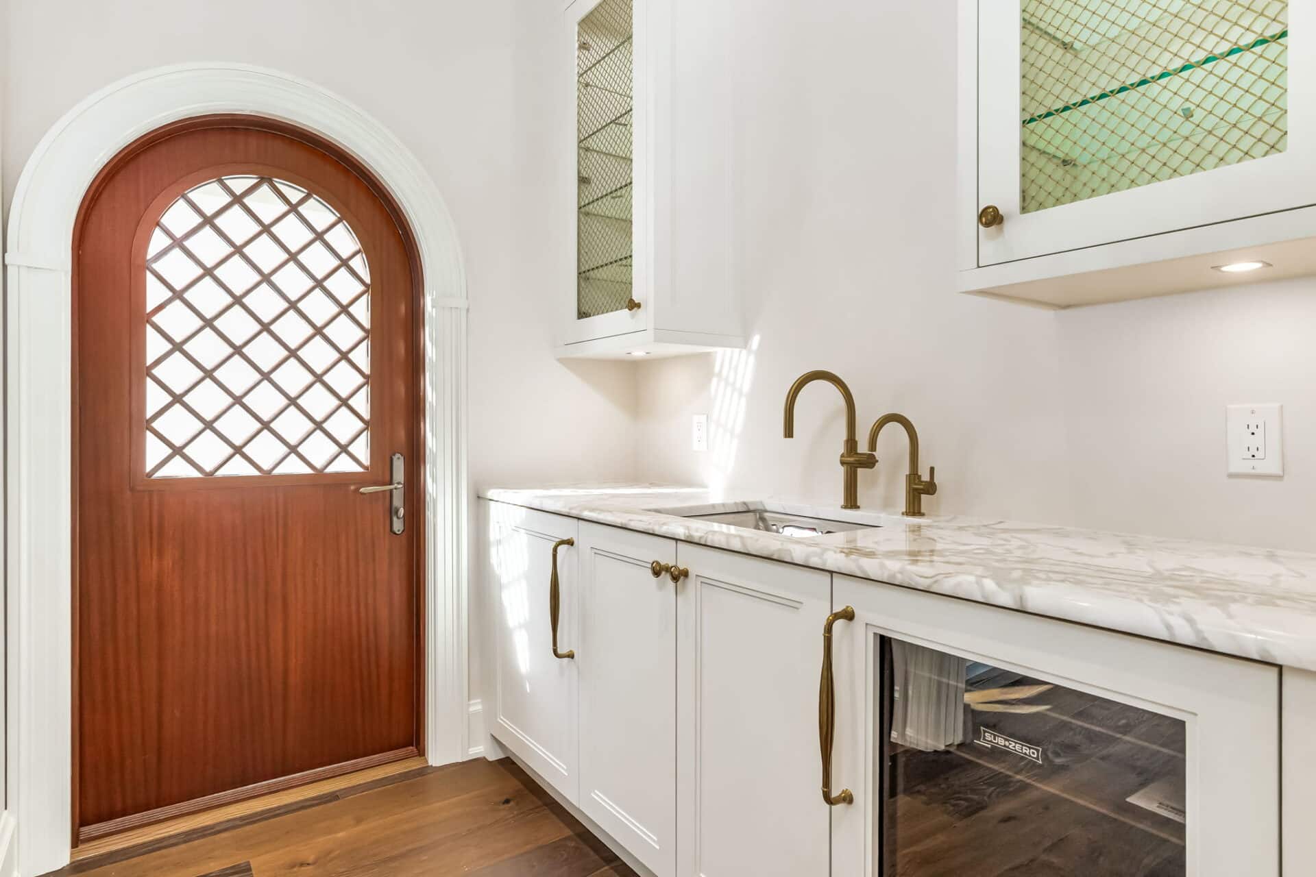 Close-up of a built-in wet bar with white cabinetry, a marble countertop, antique-brass fixtures, and a dramatic arched wooden door with a diamond grid.