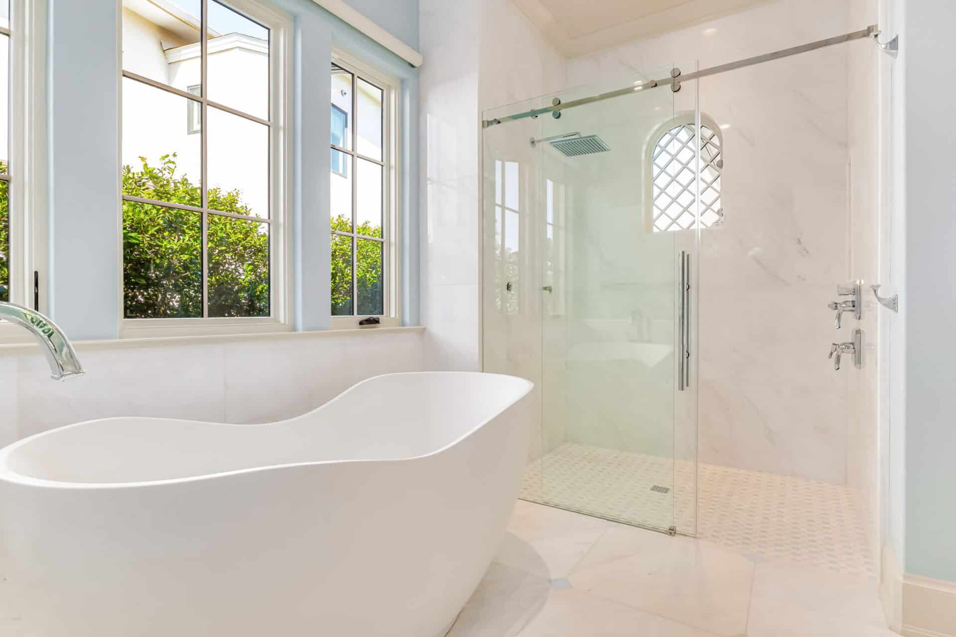 Luxury bathroom with a modern freestanding tub, marble walls and floor, a glass-enclosed shower, and large windows with privacy-grid details.