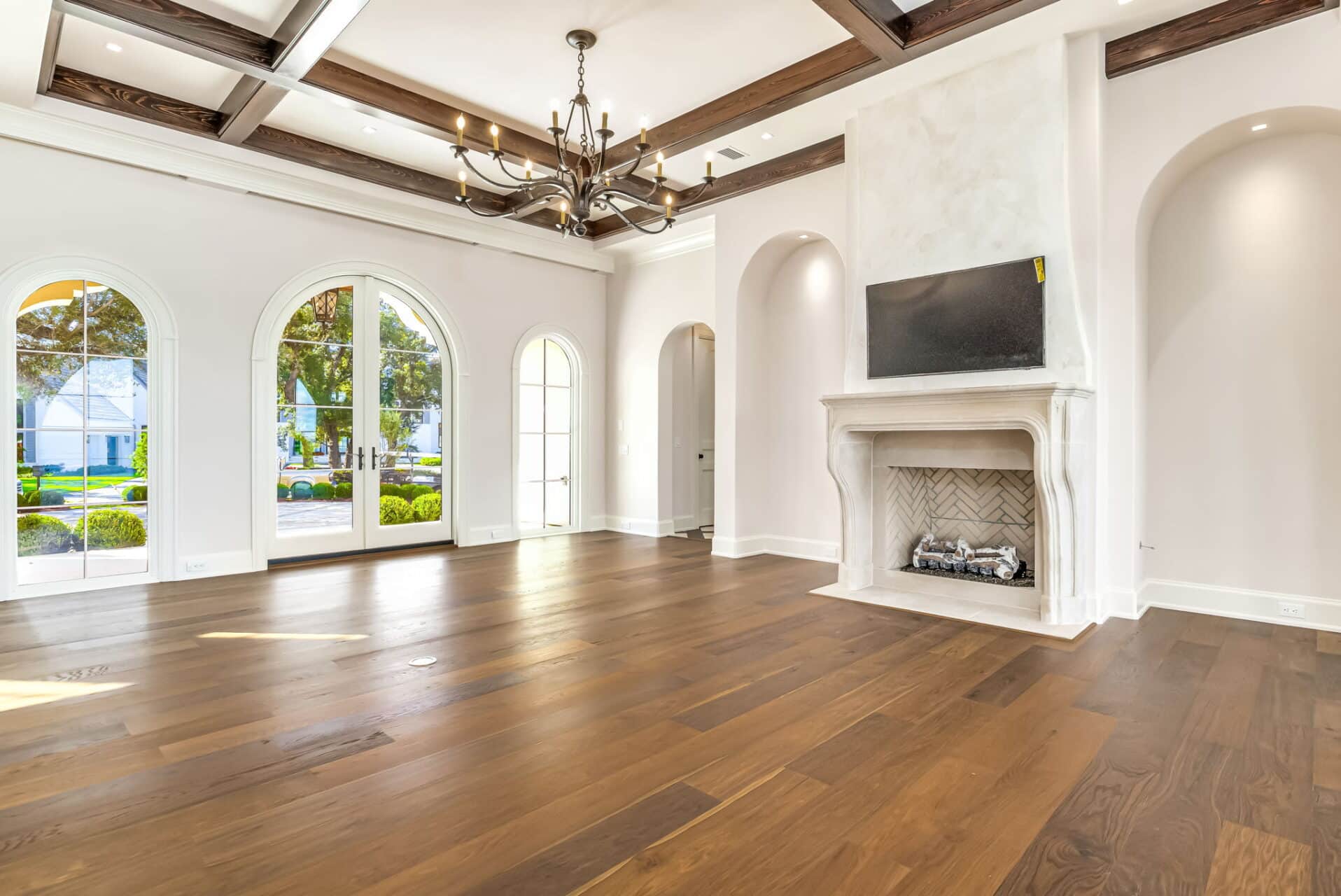 Large empty living room with wood floors, a limestone fireplace, a TV mounted above, a dark wood-beamed ceiling, and three arched doors to the outside.