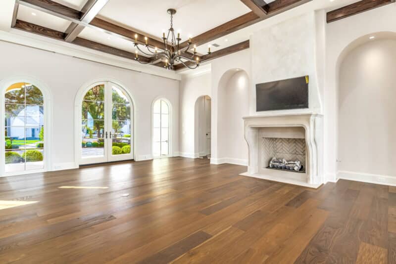Large empty living room with wood floors, a limestone fireplace, a TV mounted above, a dark wood-beamed ceiling, and three arched doors to the outside.