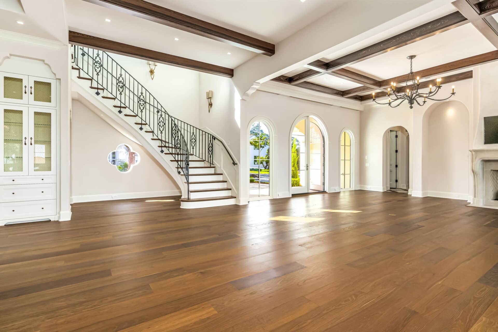 Spacious main living area with dark wood floors, a dramatic coffered ceiling, a white built-in hutch, and a grand staircase with an iron railing.