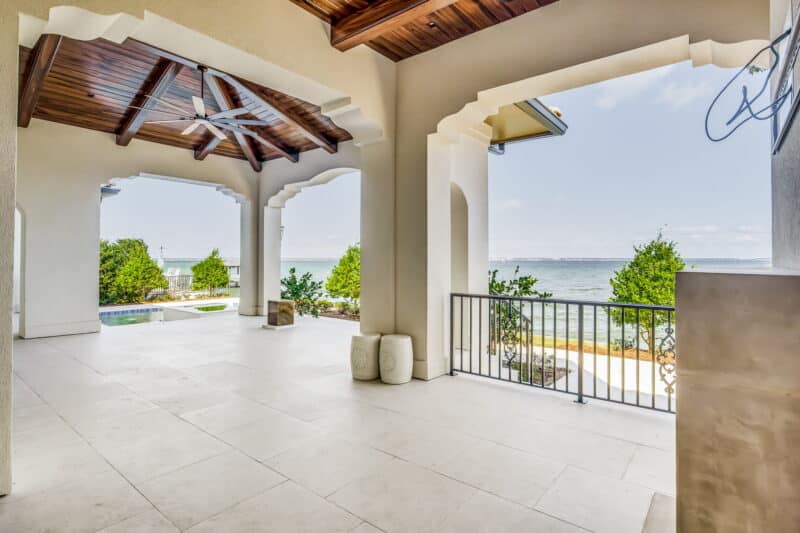 Expansive covered patio with decorative columns, a dark wood plank ceiling, a ceiling fan, and a panoramic view of the water and coastline.