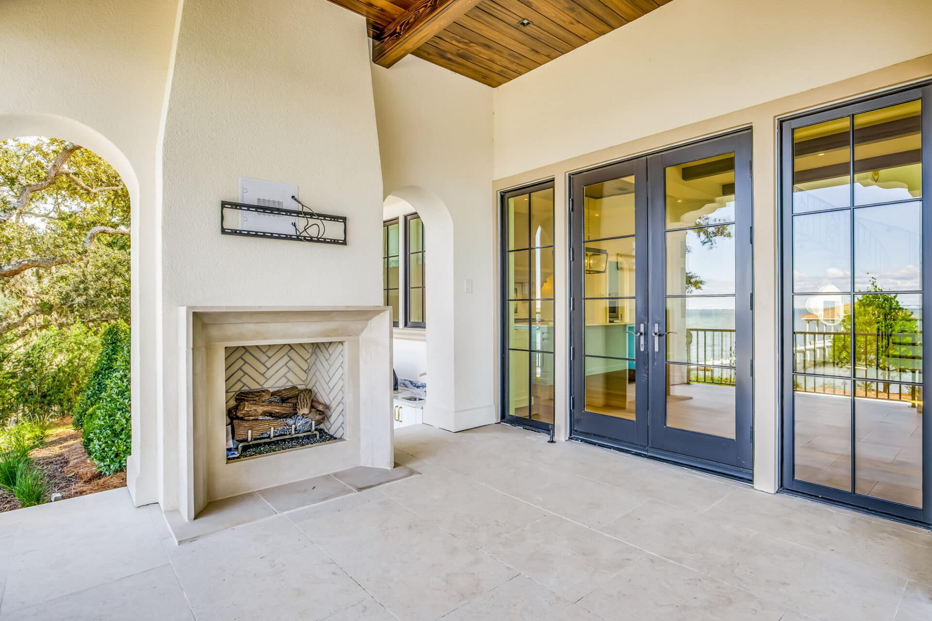 Covered patio corner featuring a white stone-clad fireplace with a herringbone-tiled interior, next to black-framed glass French doors leading indoors.