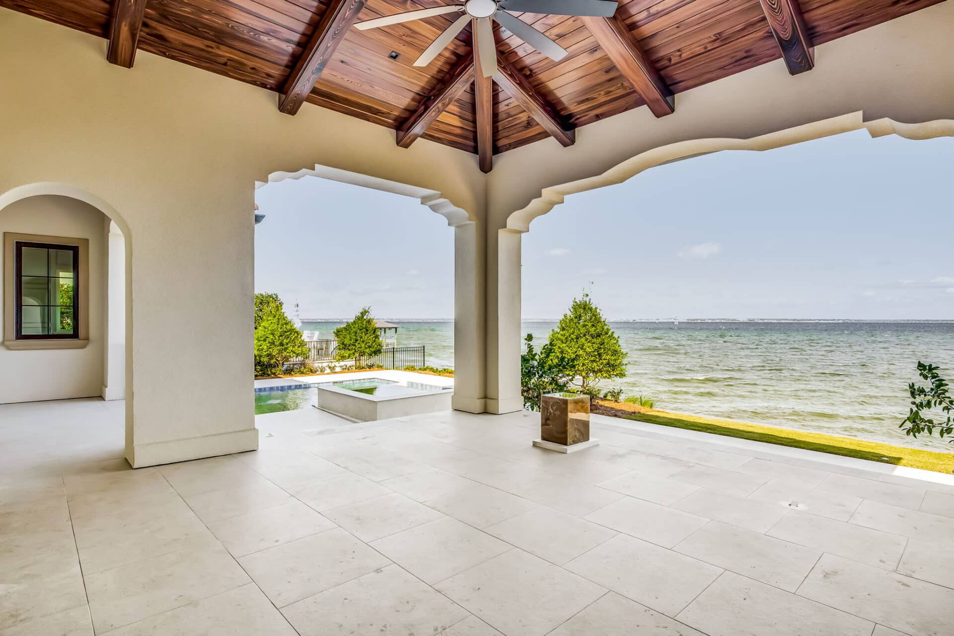 Covered patio looking out over the water, showing light-colored tiled floor, arched openings, and a modern ceiling fan in the wood-beamed ceiling.