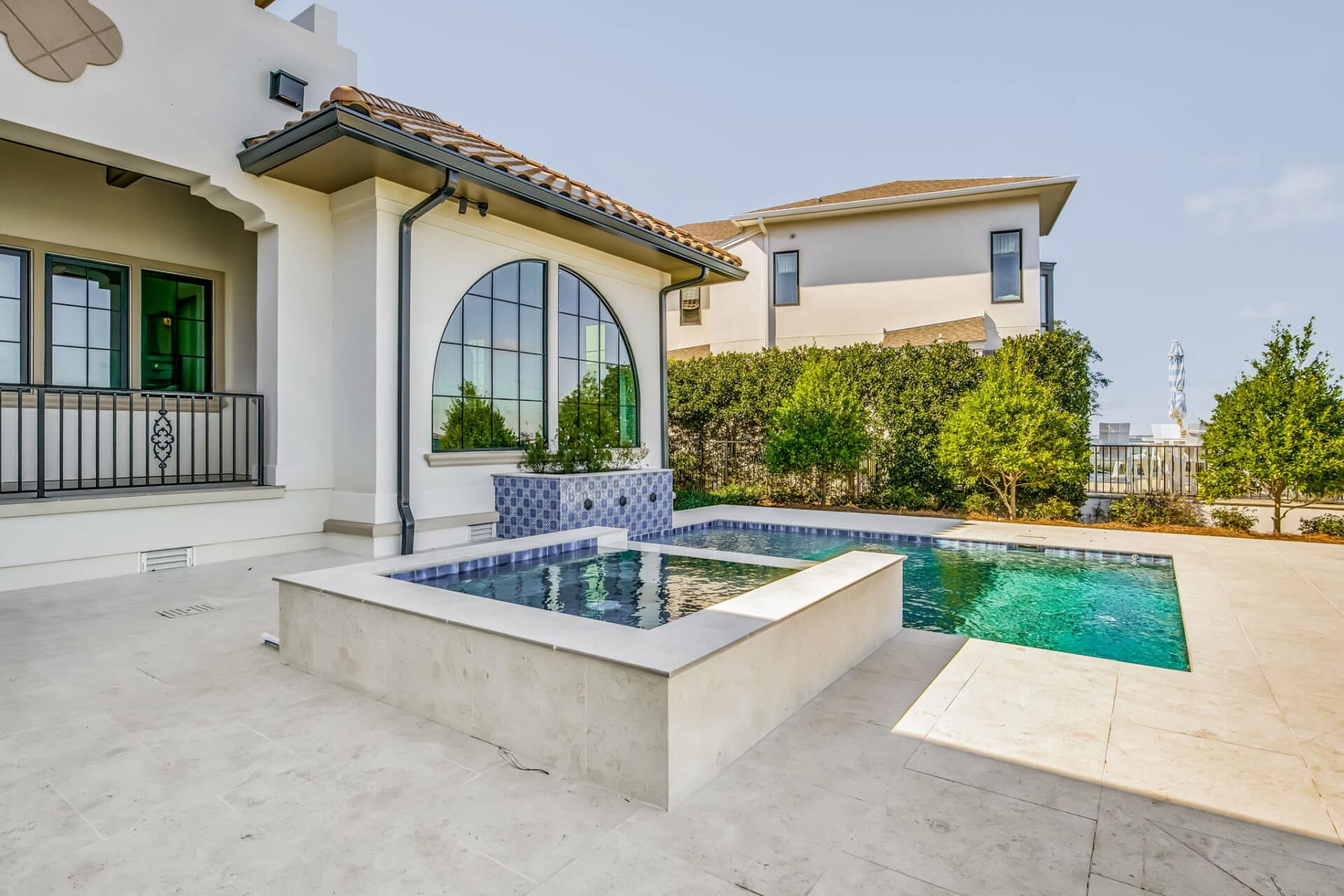 View of the swimming pool and raised spa from the house, showing the large arched window of the home and a blue sky overhead.