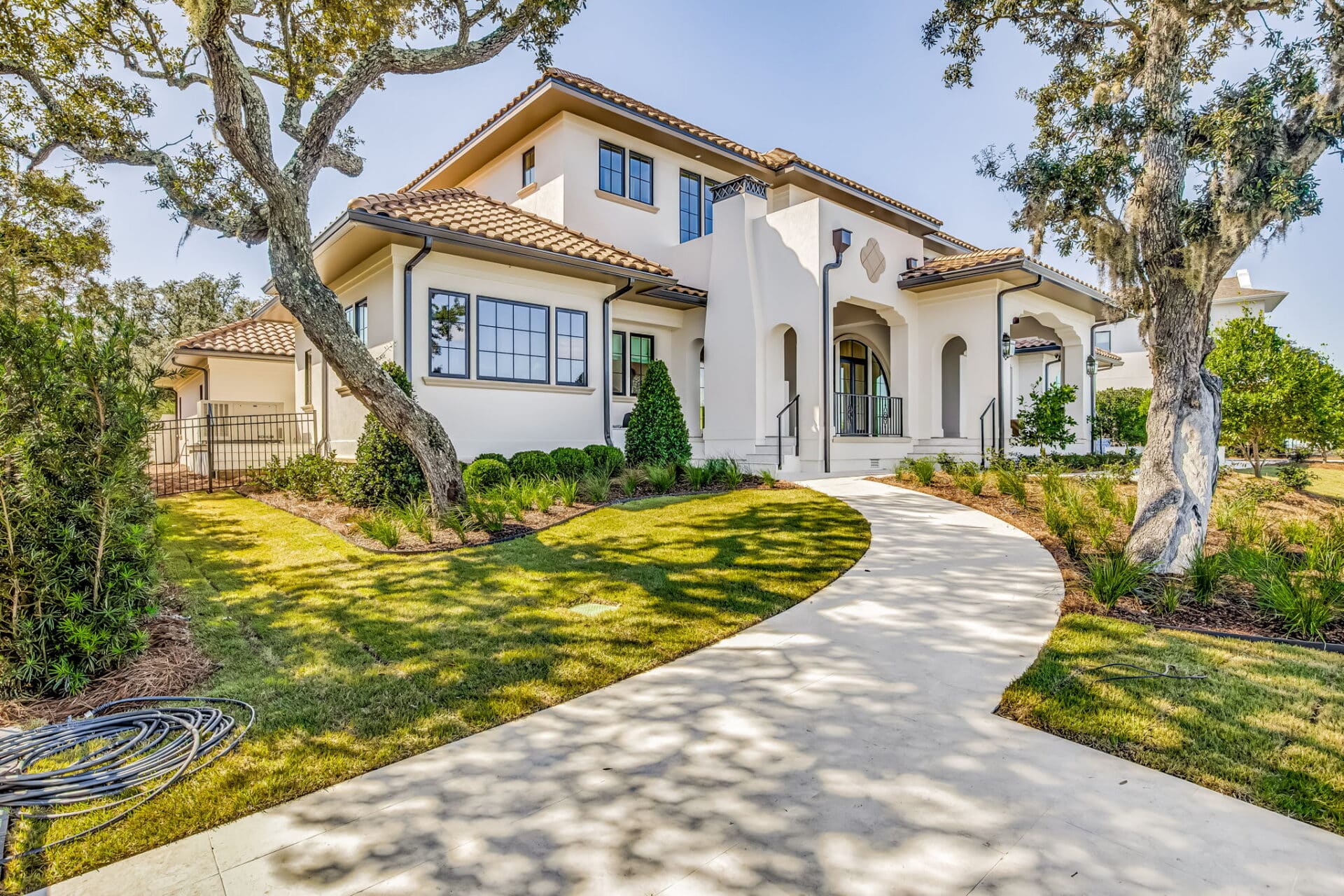 Front exterior of a stucco, two-story waterfront home with a tile roof, winding driveway, and large oak trees.