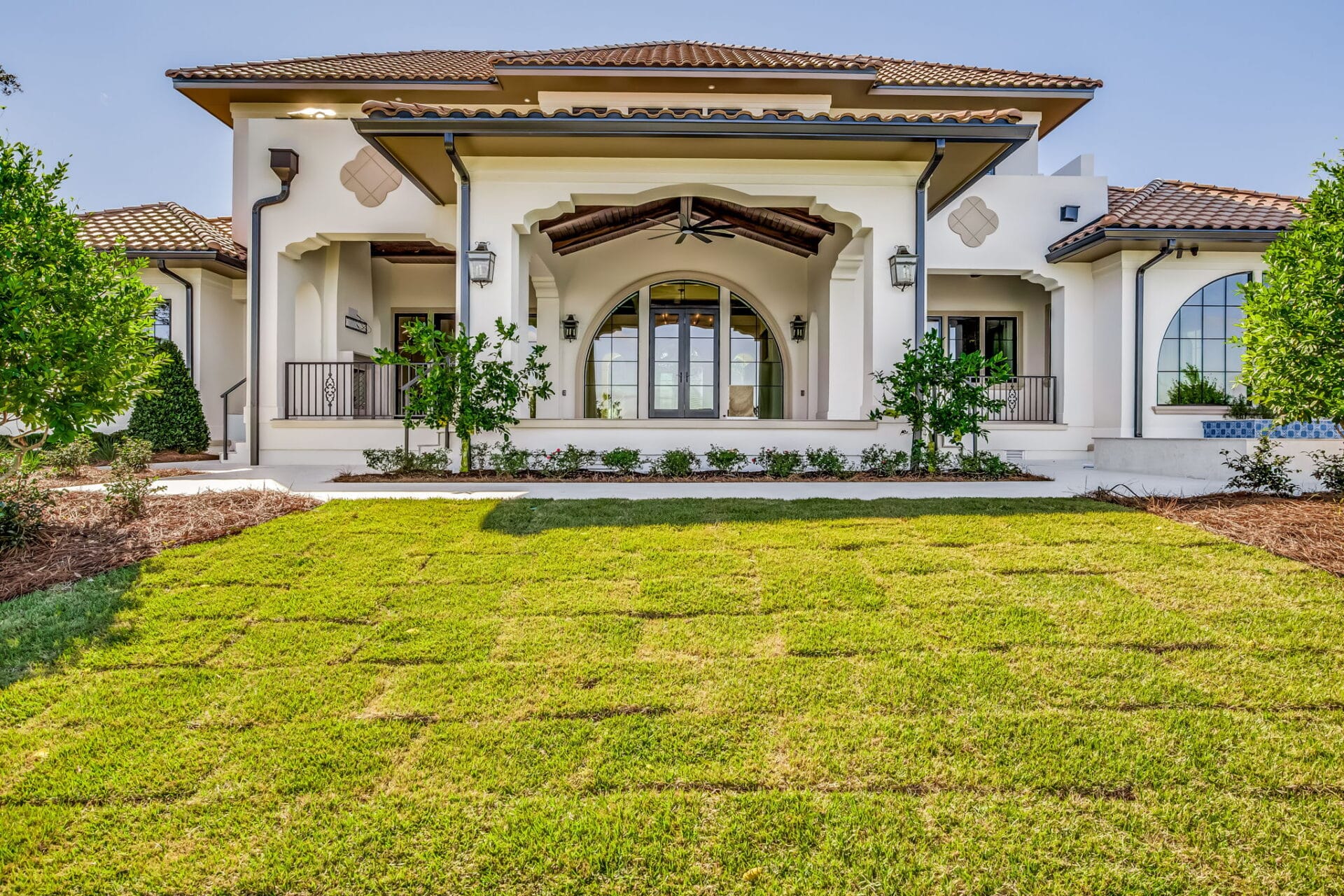 Rear facade of a luxury waterfront home overlooking a lawn, featuring a central covered terrace with a large arched window and a tile roof.