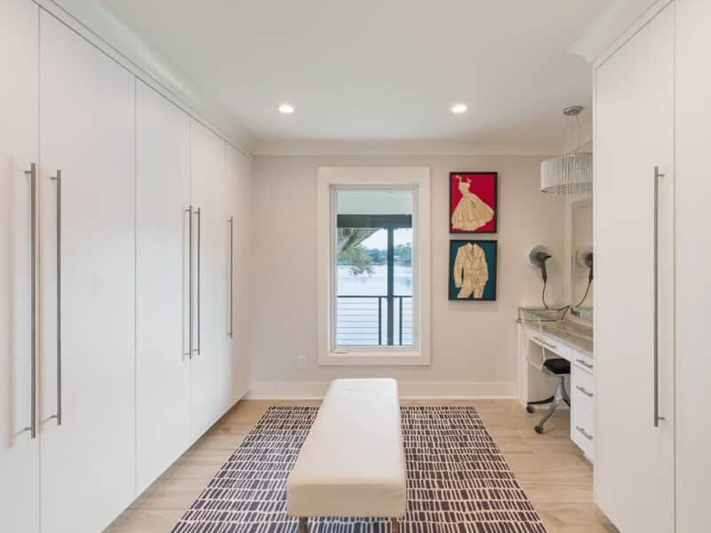 Modern, spacious white walk-in closet featuring floor-to-ceiling wardrobes, a central upholstered bench, a built-in vanity station, and a large window overlooking a scenic lake.