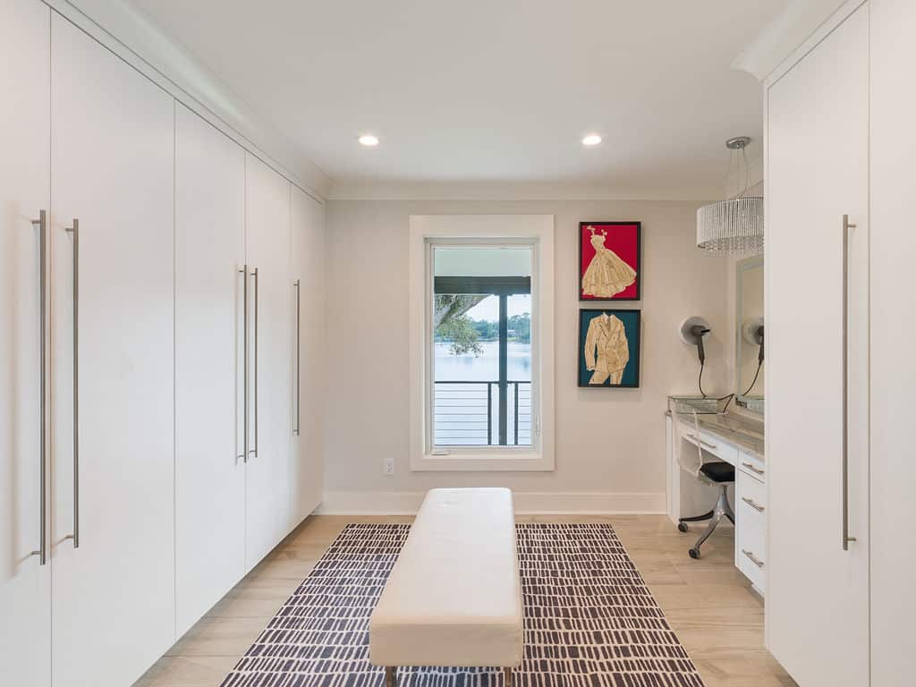 Modern, spacious white walk-in closet featuring floor-to-ceiling wardrobes, a central upholstered bench, a built-in vanity station, and a large window overlooking a scenic lake.