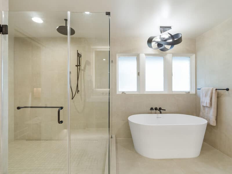 Modern luxury bathroom remodel featuring a glass-enclosed walk-in shower with matte black fixtures next to a freestanding white soaking tub beneath three windows and a contemporary ribbon light fixture.