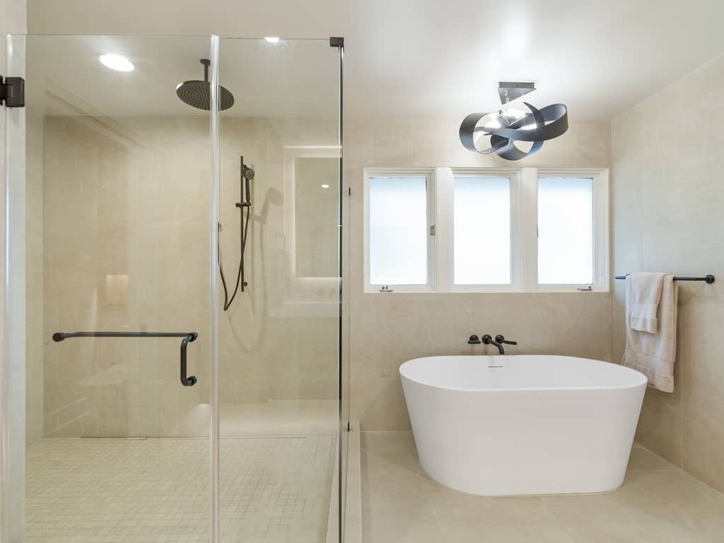Modern luxury bathroom remodel featuring a glass-enclosed walk-in shower with matte black fixtures next to a freestanding white soaking tub beneath three windows and a contemporary ribbon light fixture.