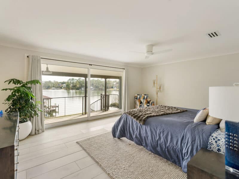 Modern bedroom with light wood flooring, a blue-clothed bed, and large sliding glass doors opening to a scenic waterfront balcony and boat dock.