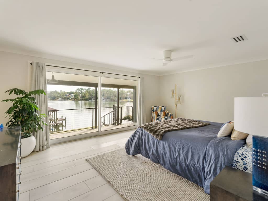 Modern bedroom with light wood flooring, a blue-clothed bed, and large sliding glass doors opening to a scenic waterfront balcony and boat dock.