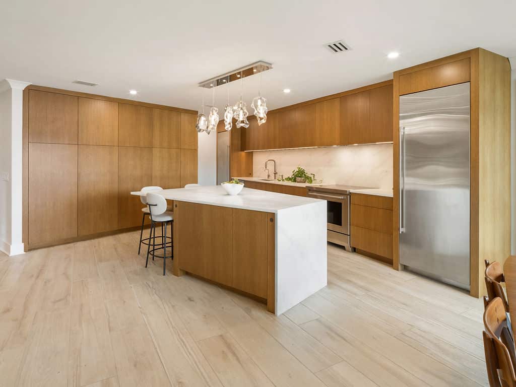 Large modern open-concept kitchen with custom white oak cabinets, a waterfall marble island with bar stools, stainless steel appliances, and a unique glass pendant light fixture.