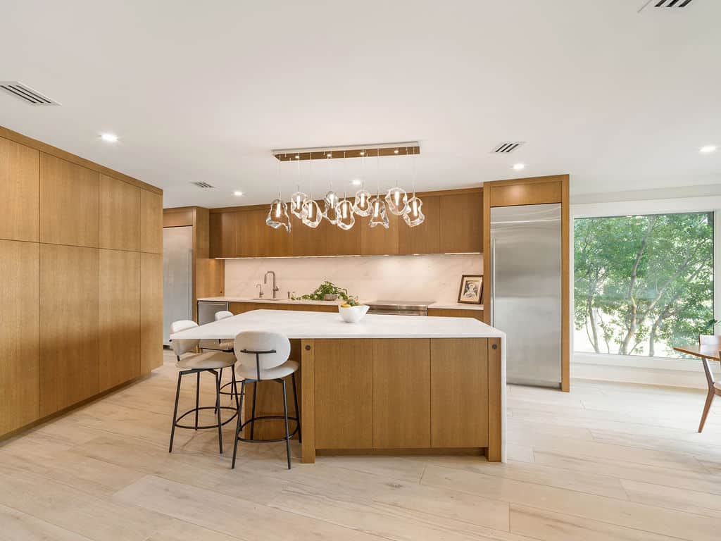 Modern minimalist kitchen featuring light wood cabinetry, a large white marble island with bar stools, stainless steel appliances, and unique glass pendant lights.