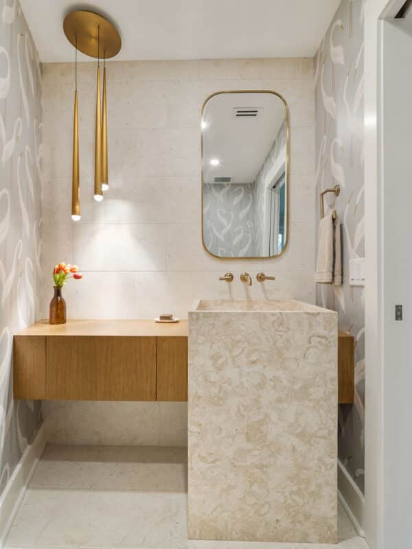 Modern powder room featuring a floor-to-ceiling cream marble sink, floating wooden vanity, gold pendant lights, and gray swan-patterned wallpaper.