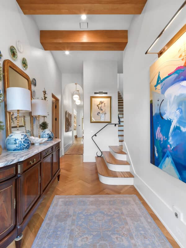 Bright entryway featuring herringbone wood flooring, large abstract blue wall art, a vintage wooden credenza with marble top, and a staircase leading upstairs.
