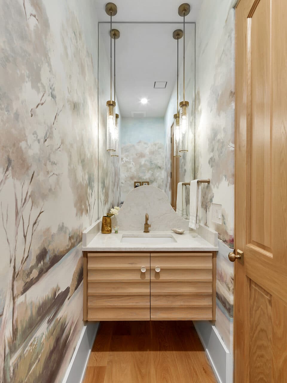 Luxury powder room featuring a custom landscape wall mural, a floating light oak vanity with a fluted facade, an arched marble backsplash, and hanging cylinder pendant lights.