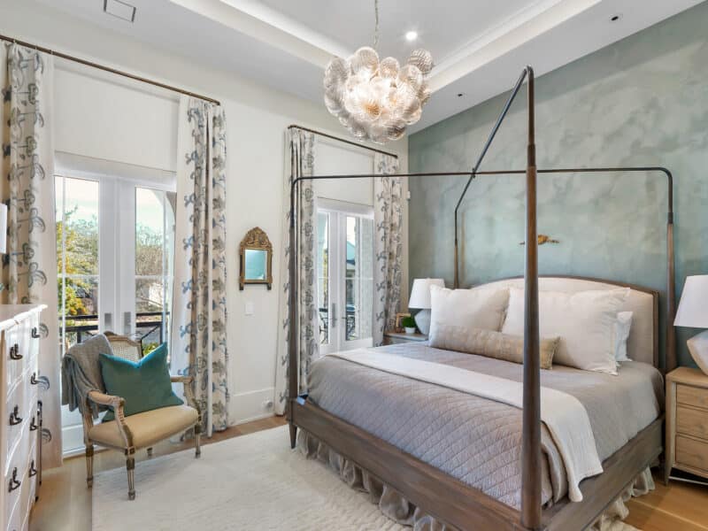 A primary bedroom with a dark wood four-poster bed frame, textured seafoam green accent wall, crystal bubble chandelier, and French doors leading to a balcony.