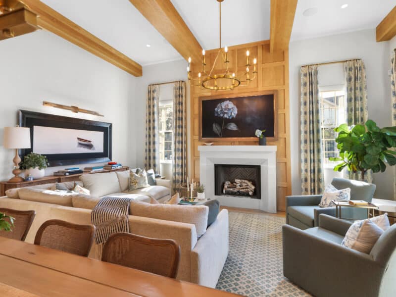 Spacious living room with high ceilings, exposed wood beams, a light sectional sofa, two green armchairs, a modern white fireplace, and a wood-paneled accent wall with large floral artwork.