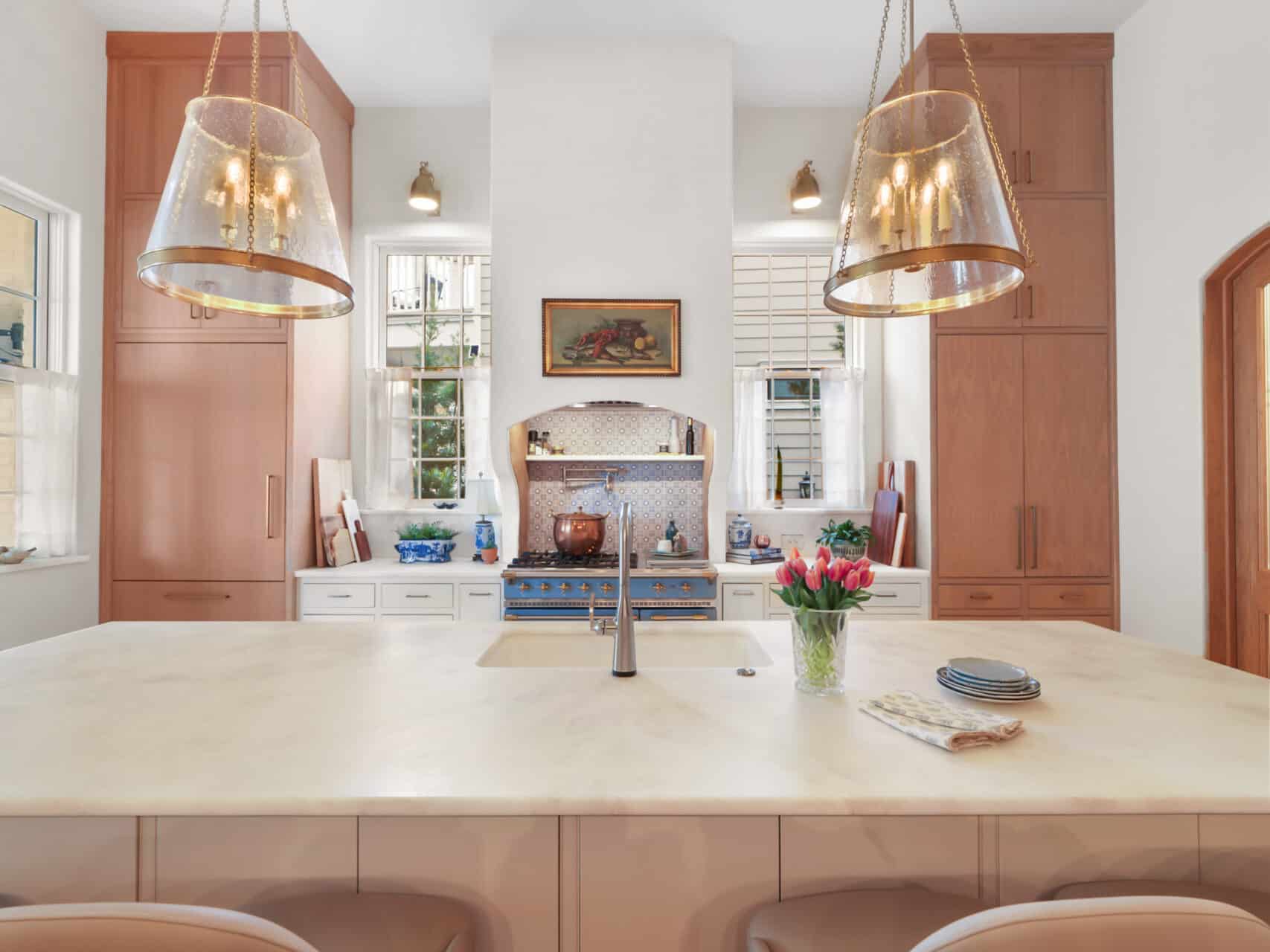 Bright luxury kitchen with a massive white marble island, light wood cabinetry, a custom white range hood over a blue French-style gas stove, and two oversized brass and glass pendant lights.