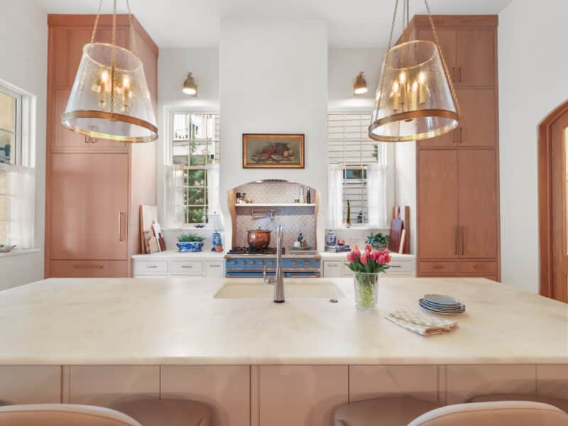 Bright luxury kitchen with a massive white marble island, light wood cabinetry, a custom white range hood over a blue French-style gas stove, and two oversized brass and glass pendant lights.
