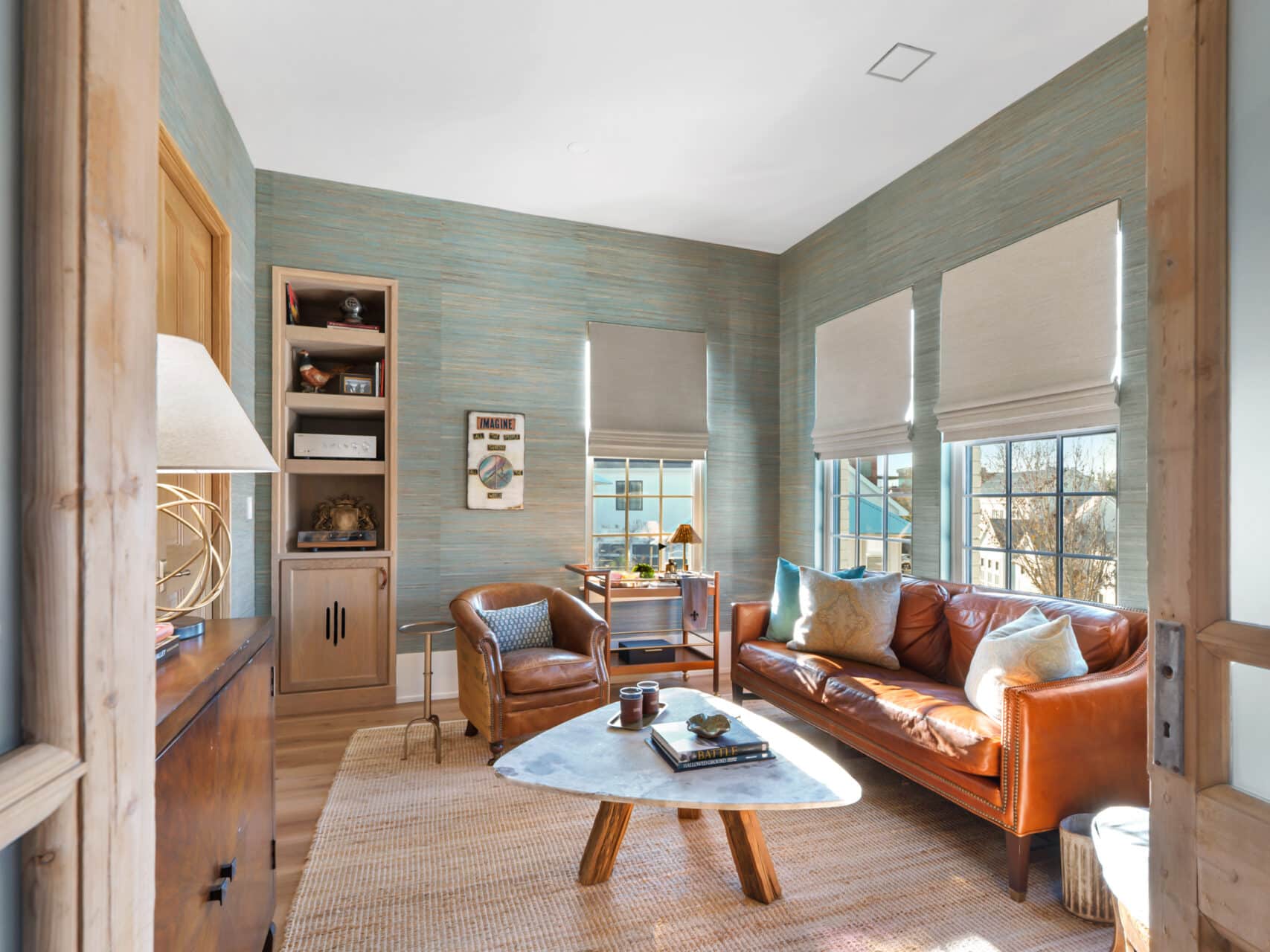 Cozy den with textured teal grass cloth wallpaper, a tan leather sofa, a triangular marble coffee table, and natural wood built-in shelving.
