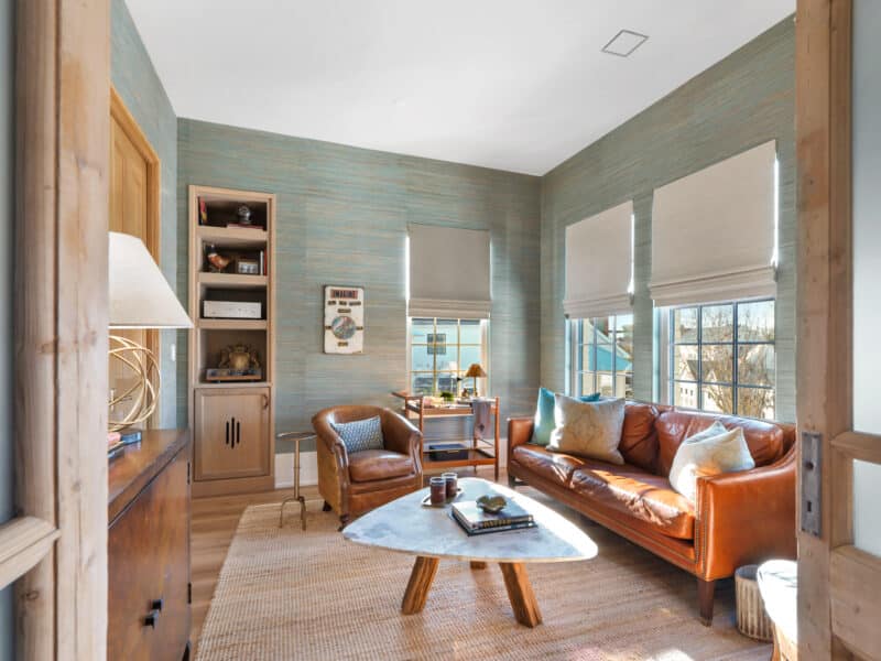 Cozy den with textured teal grass cloth wallpaper, a tan leather sofa, a triangular marble coffee table, and natural wood built-in shelving.