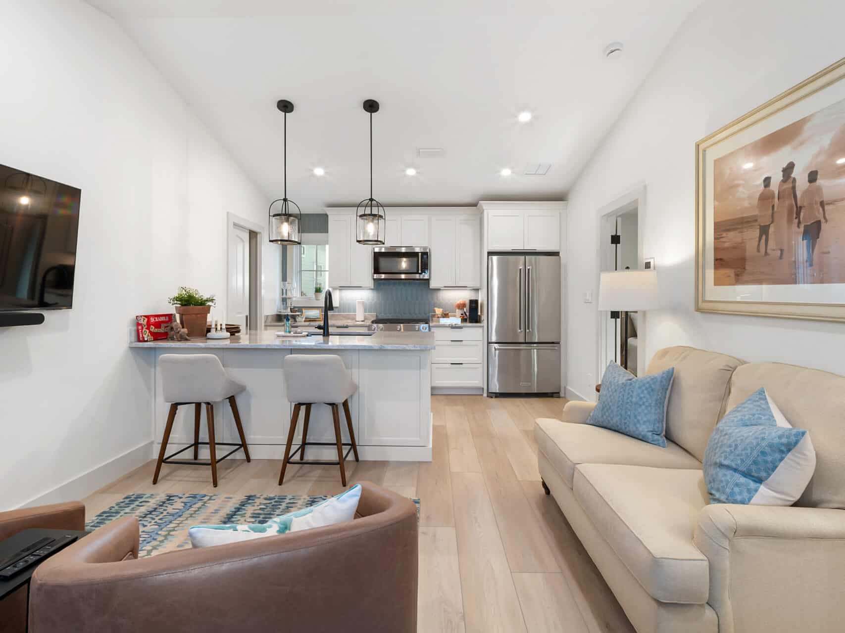 Open-concept living area and kitchen featuring light hardwood floors, a neutral sofa with blue accent pillows, a marble-top kitchen island with barstools, and white shaker cabinets with stainless steel appliances.