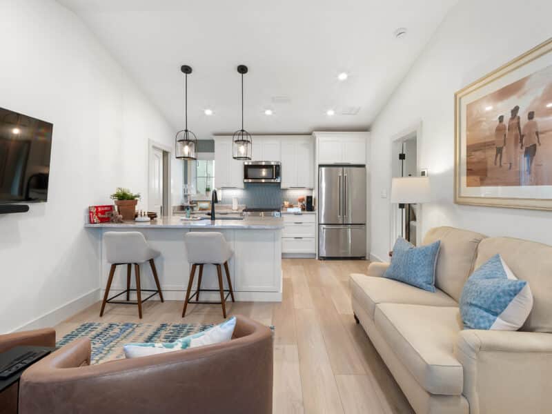 Open-concept living area and kitchen featuring light hardwood floors, a neutral sofa with blue accent pillows, a marble-top kitchen island with barstools, and white shaker cabinets with stainless steel appliances.
