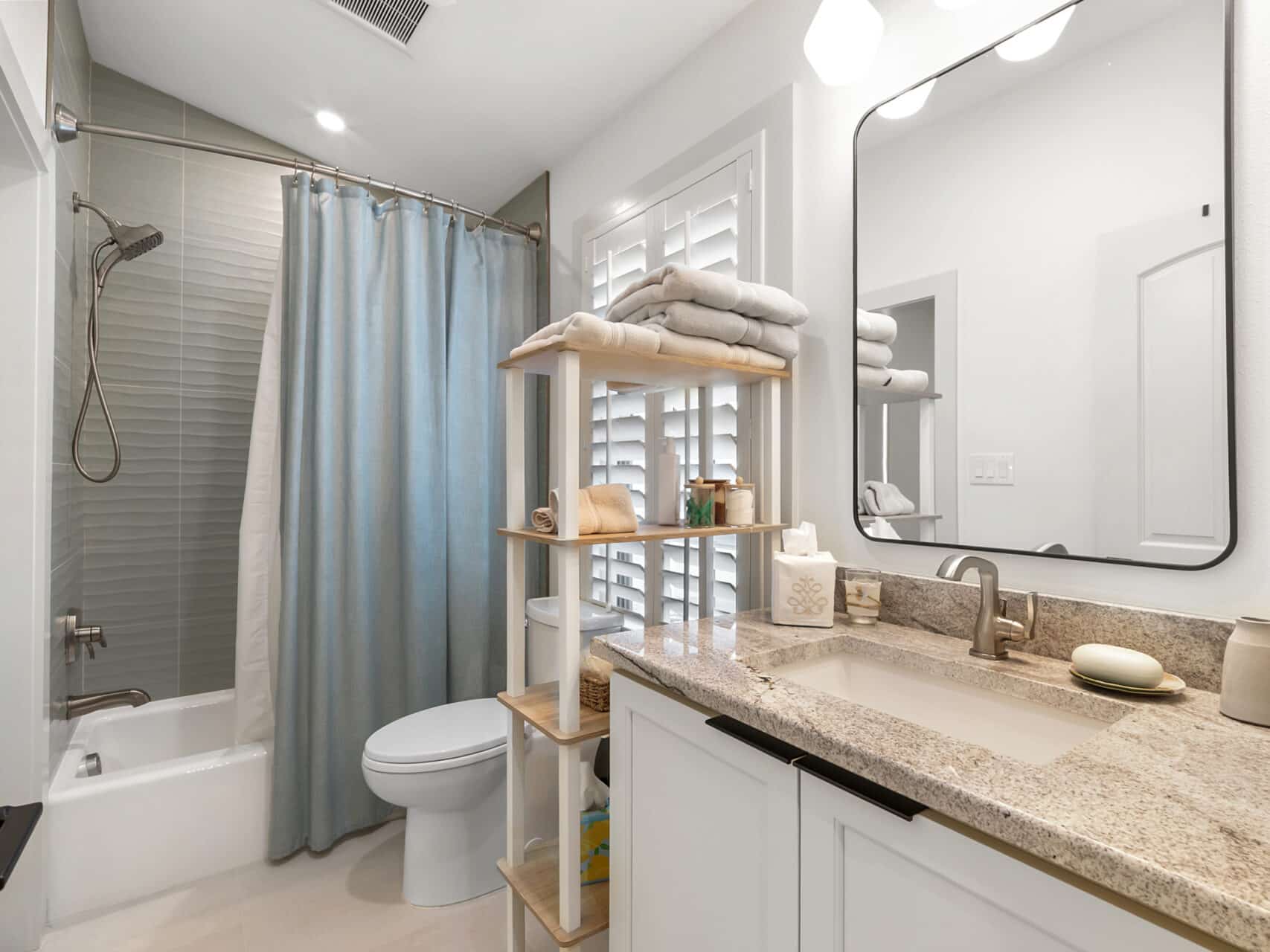 A clean bathroom featuring a white vanity with a granite countertop, an open wooden shelving unit, a toilet, and a shower-tub combo with a dusty blue curtain.