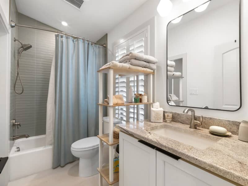 A clean bathroom featuring a white vanity with a granite countertop, an open wooden shelving unit, a toilet, and a shower-tub combo with a dusty blue curtain.