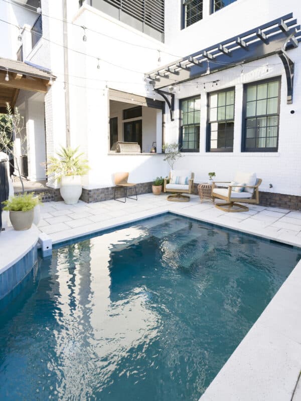 Backyard luxury pool surrounded by light stone pavers, comfortable outdoor lounge chairs, string lights, and an outdoor kitchen pass-through window.