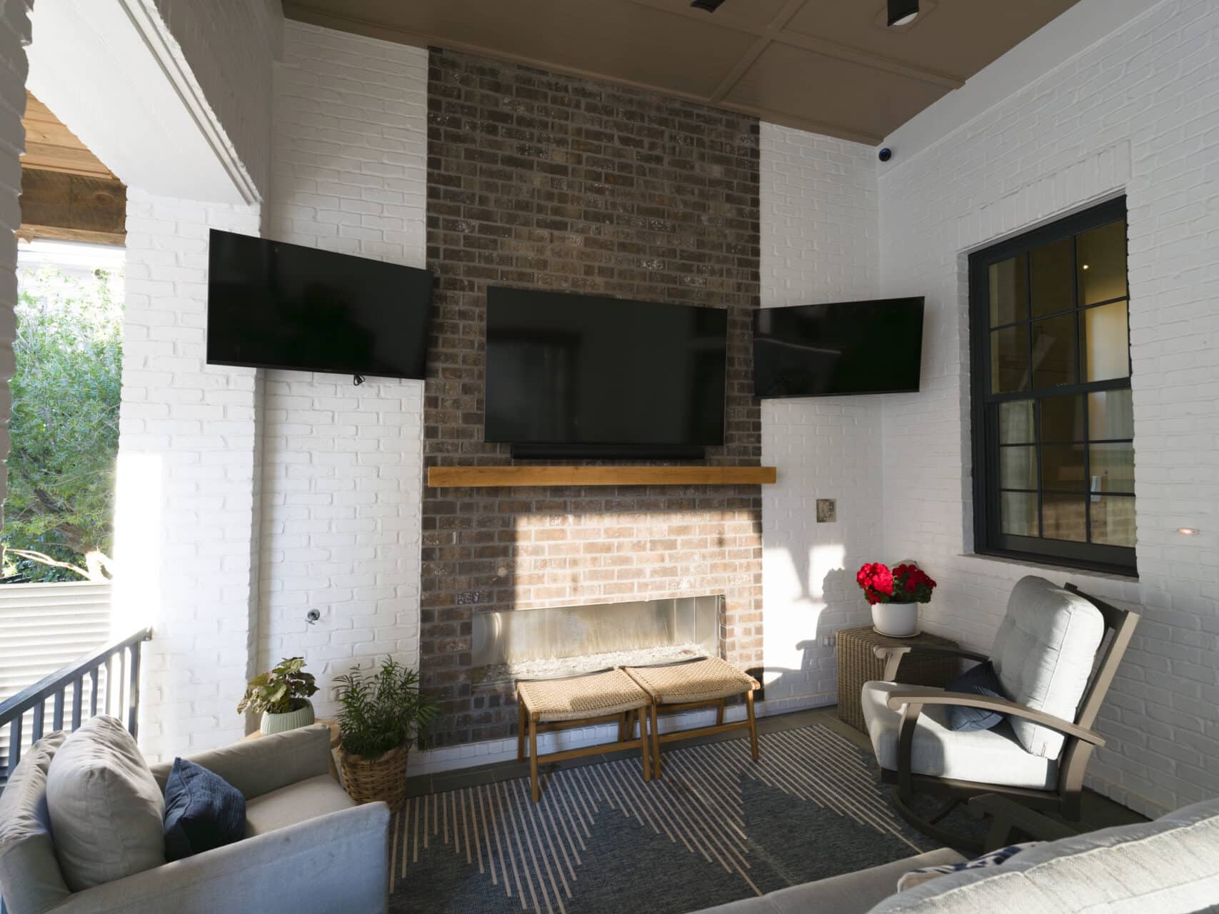 Covered outdoor patio with a floor-to-ceiling brick fireplace, a large flat-screen TV mounted above a wooden mantel, flanked by two smaller TVs on white brick walls with comfortable patio seating.