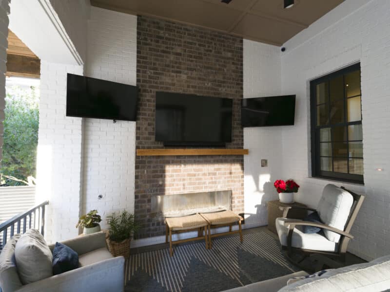 Covered outdoor patio with a floor-to-ceiling brick fireplace, a large flat-screen TV mounted above a wooden mantel, flanked by two smaller TVs on white brick walls with comfortable patio seating.