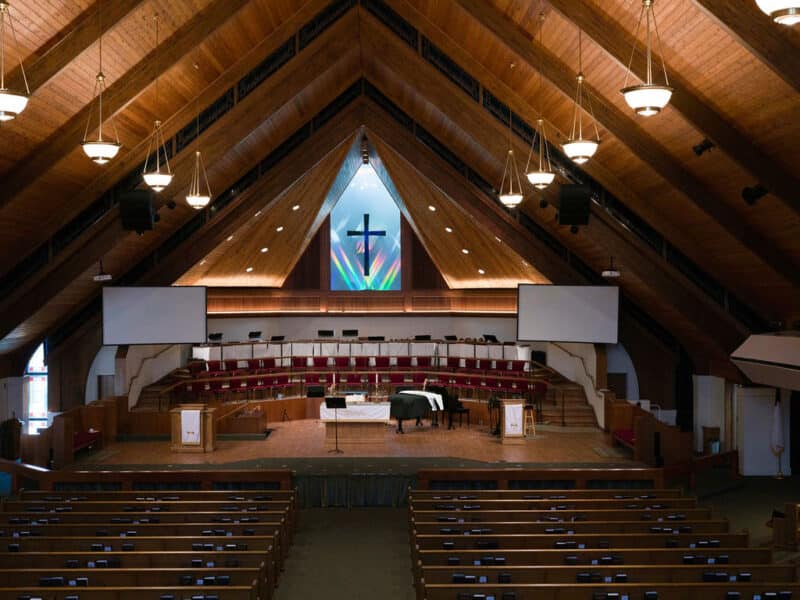 High-angle, symmetrical shot of a church altar and choir loft centered under an illuminated stained-glass style cross with hanging light fixtures.
