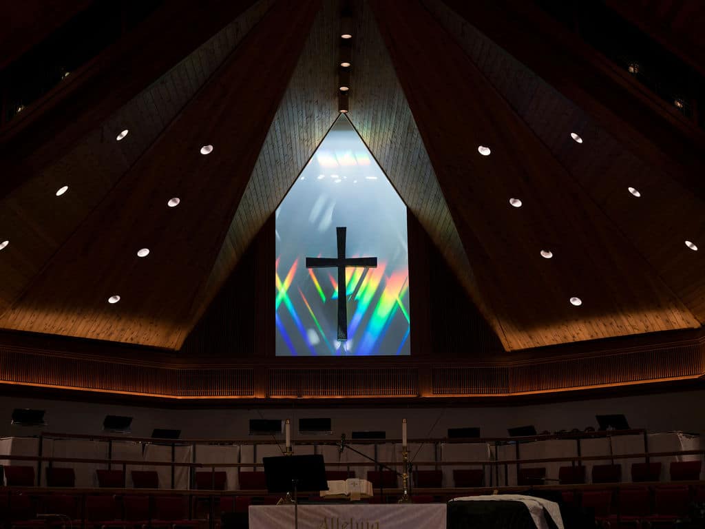 Close-up of a Christian cross beneath a vaulted wooden ceiling that is illuminated by a prism that refracts the sun.