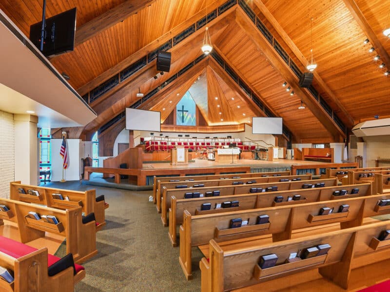 Side-angle interior view of a modern church featuring rows of wooden pews, a multi-tiered altar stage, and a natural wood-paneled vaulted ceiling.