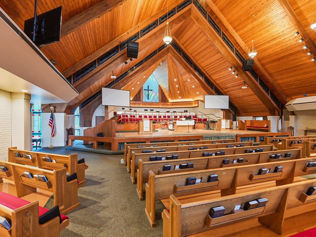 Side-angle interior view of a modern church featuring rows of wooden pews, a multi-tiered altar stage, and a natural wood-paneled vaulted ceiling.