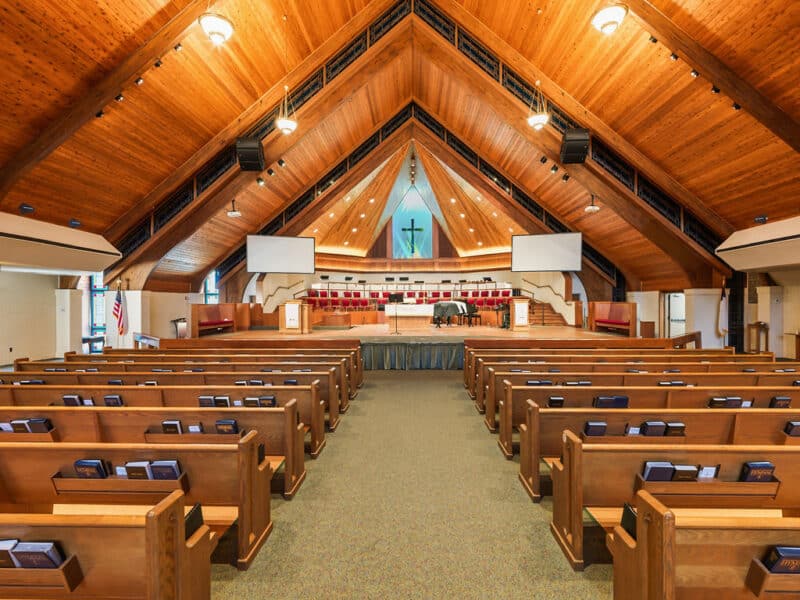 A symmetrical, wide-angle view from the center aisle of a large church with rows of wooden pews leading to a brightly lit altar under a massive vaulted wooden ceiling.