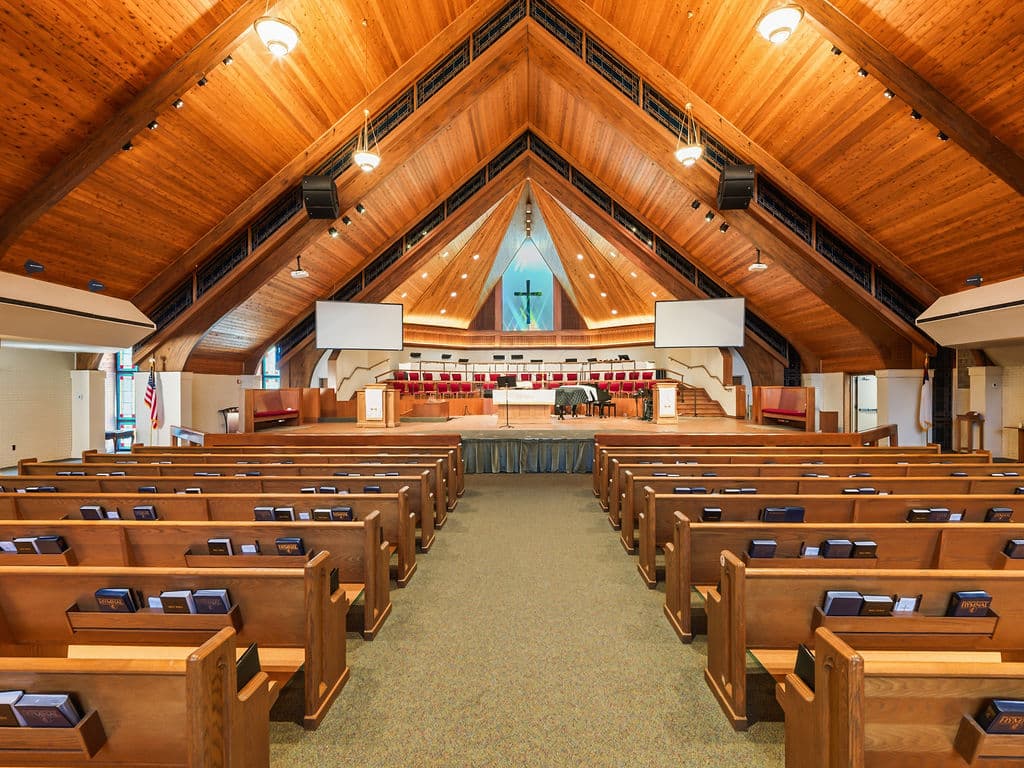 A symmetrical, wide-angle view from the center aisle of a large church with rows of wooden pews leading to a brightly lit altar under a massive vaulted wooden ceiling.