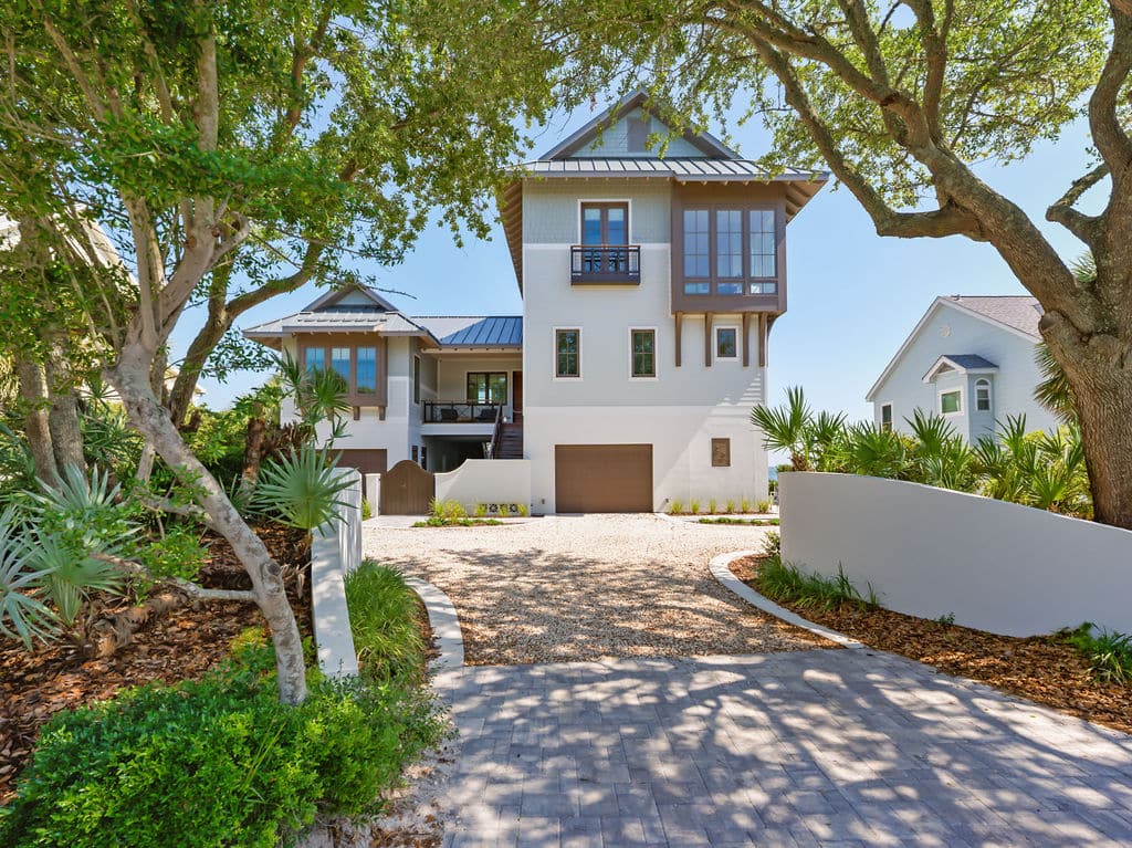 Three-story modern coastal home with light gray siding, brown trim, a gravel driveway, and lush green trees and palms.